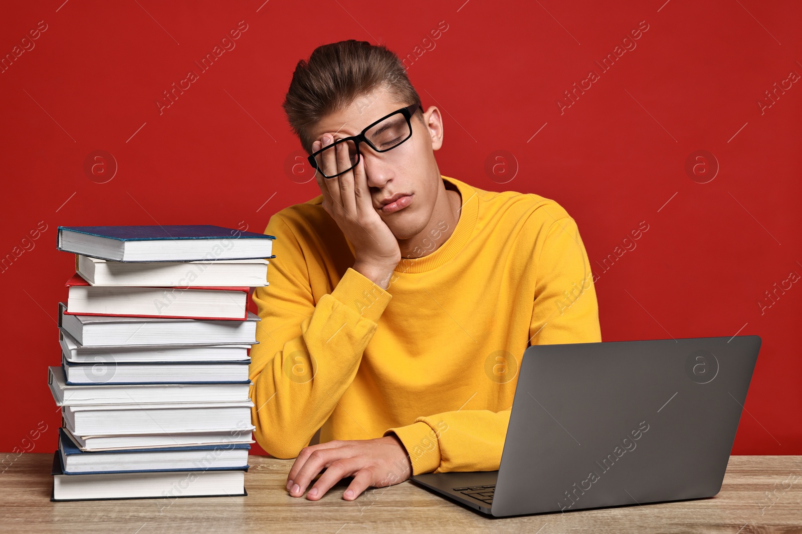 Photo of Tired student before exam with stack of books and laptop at wooden table against red background