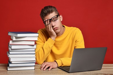 Photo of Tired student before exam with stack of books and laptop at wooden table against red background