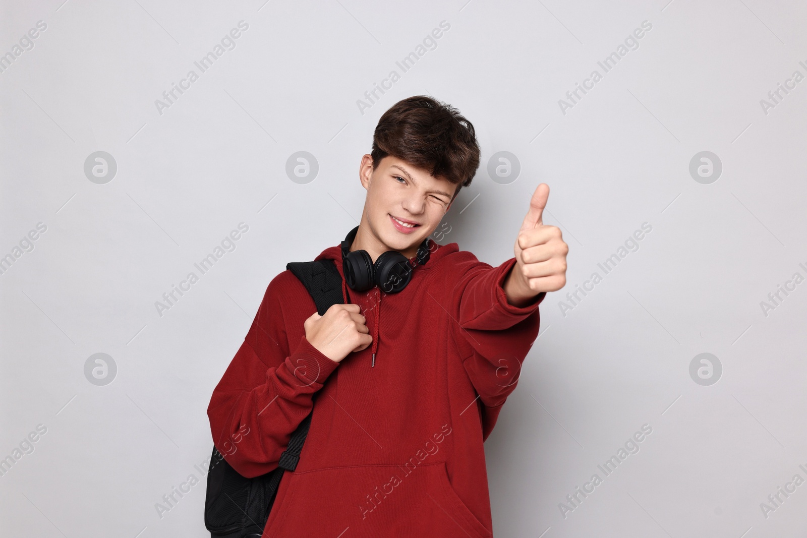 Photo of Happy teenage boy with headphones and backpack showing thumbs up on light grey background