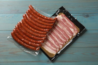 Photo of Packs of dry smoked sausages and sliced bacon on light blue wooden table, top view