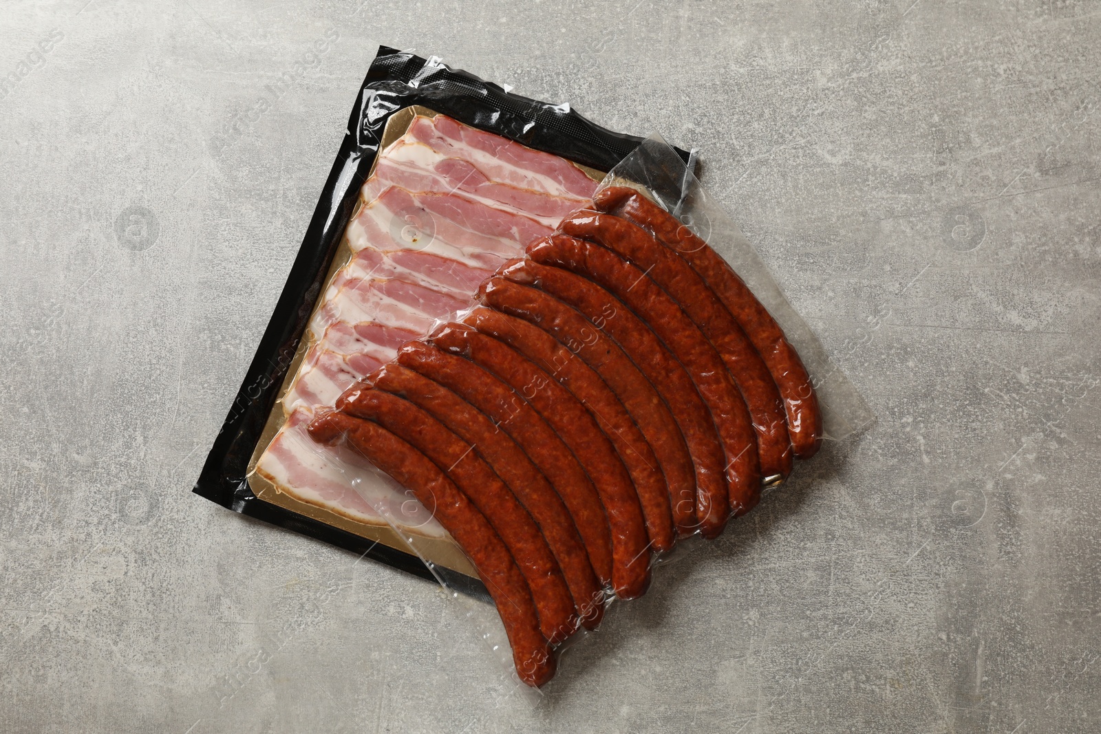 Photo of Packs of dry smoked sausages and sliced bacon on grey table, top view