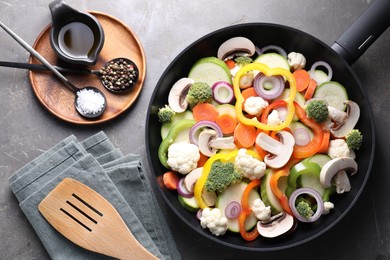 Frying pan with mix of vegetables, mushrooms, spices and slotted turner on grey table, flat lay