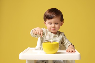 Photo of Cute little kid eating healthy baby food from bowl in high chair on yellow background