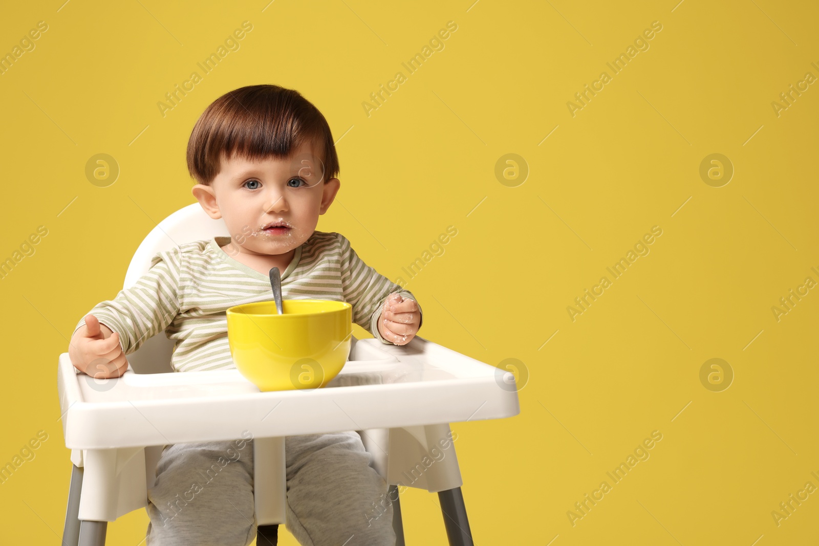 Photo of Cute little kid eating healthy baby food from bowl in high chair on yellow background, space for text