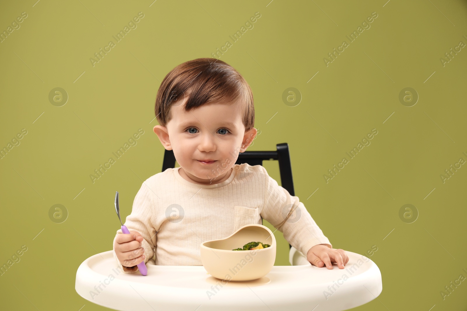 Photo of Cute little baby eating healthy food from bowl in high chair on olive background