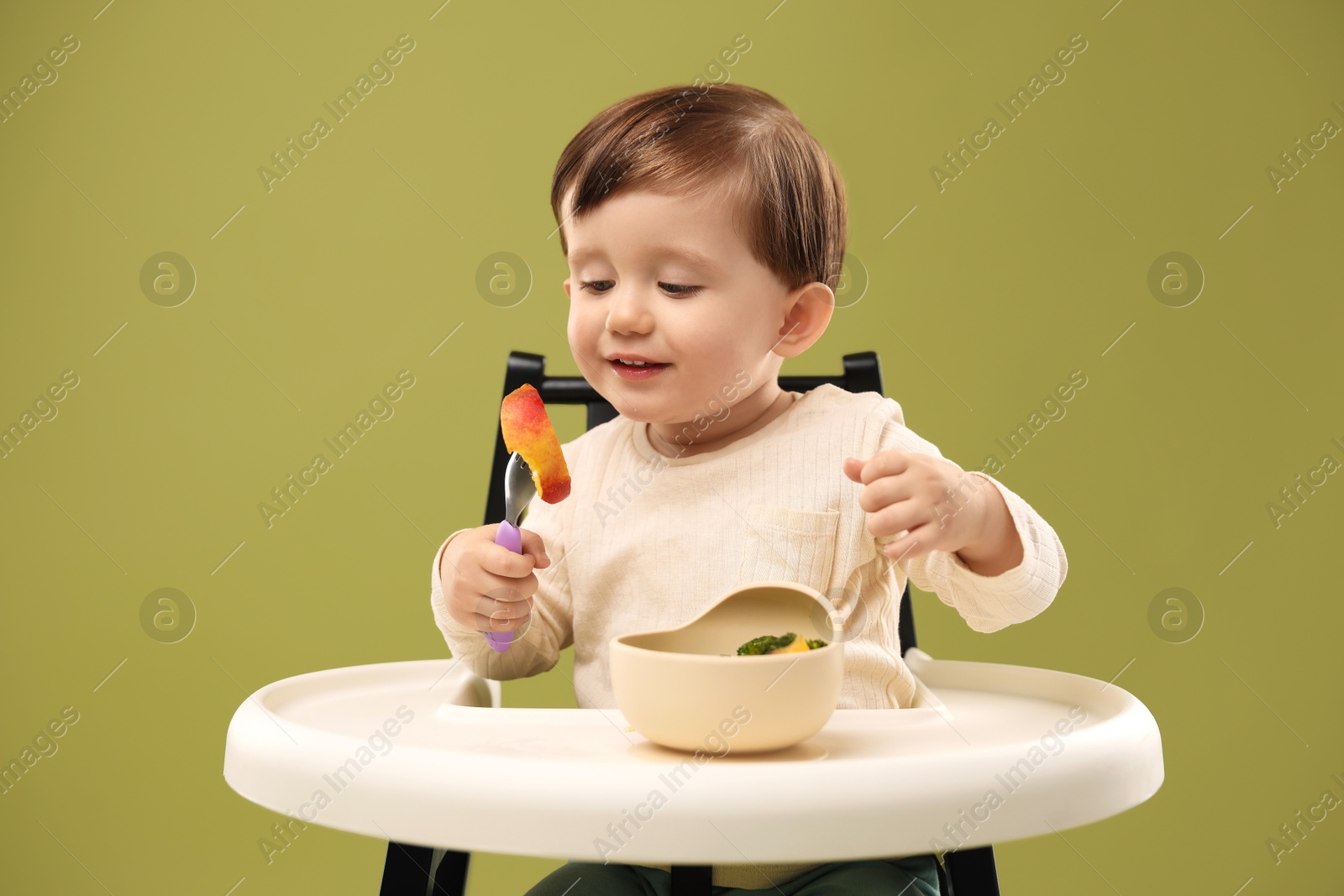 Photo of Cute little baby eating healthy food from bowl in high chair on olive background