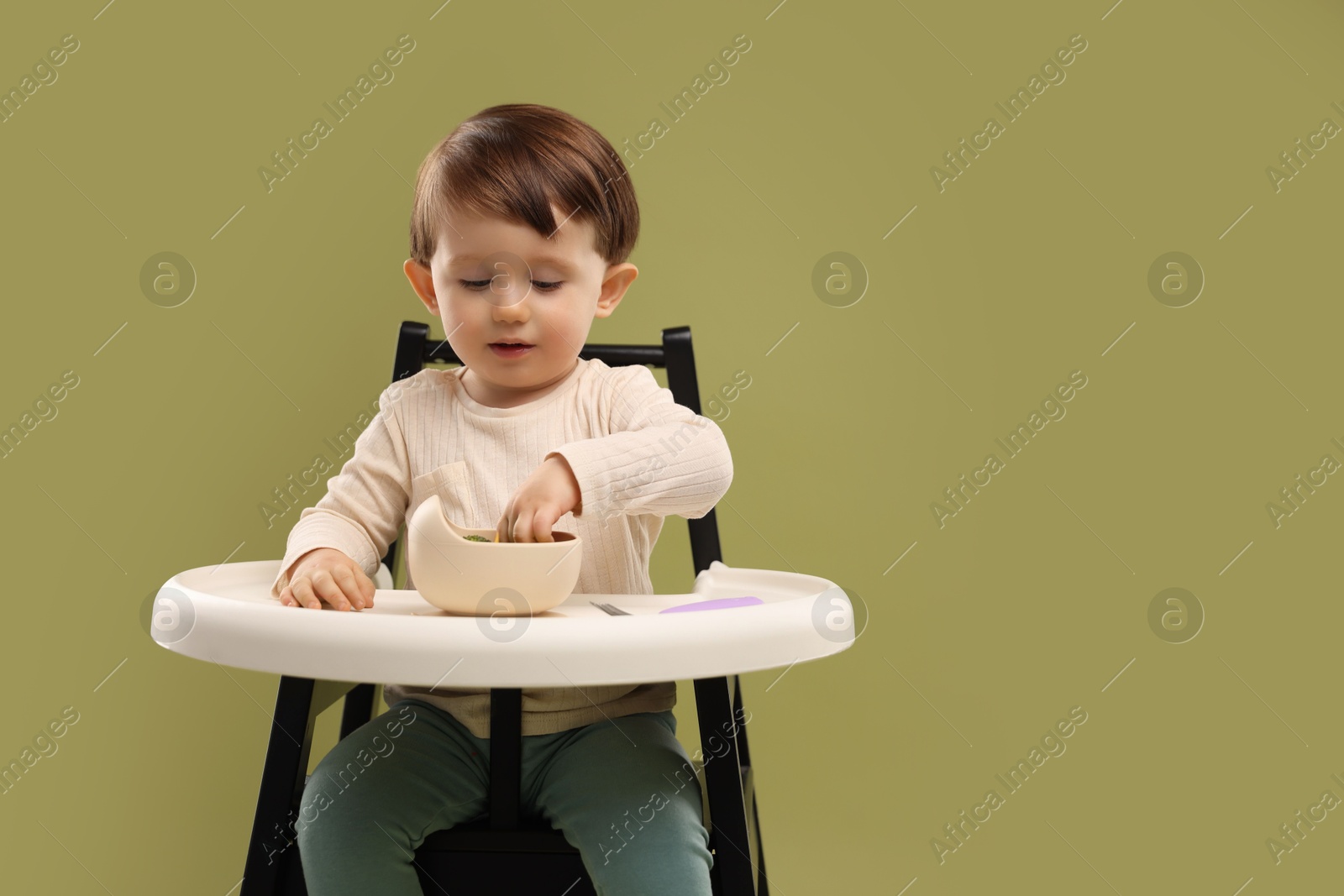 Photo of Cute little baby eating healthy food from bowl in high chair on olive background, space for text