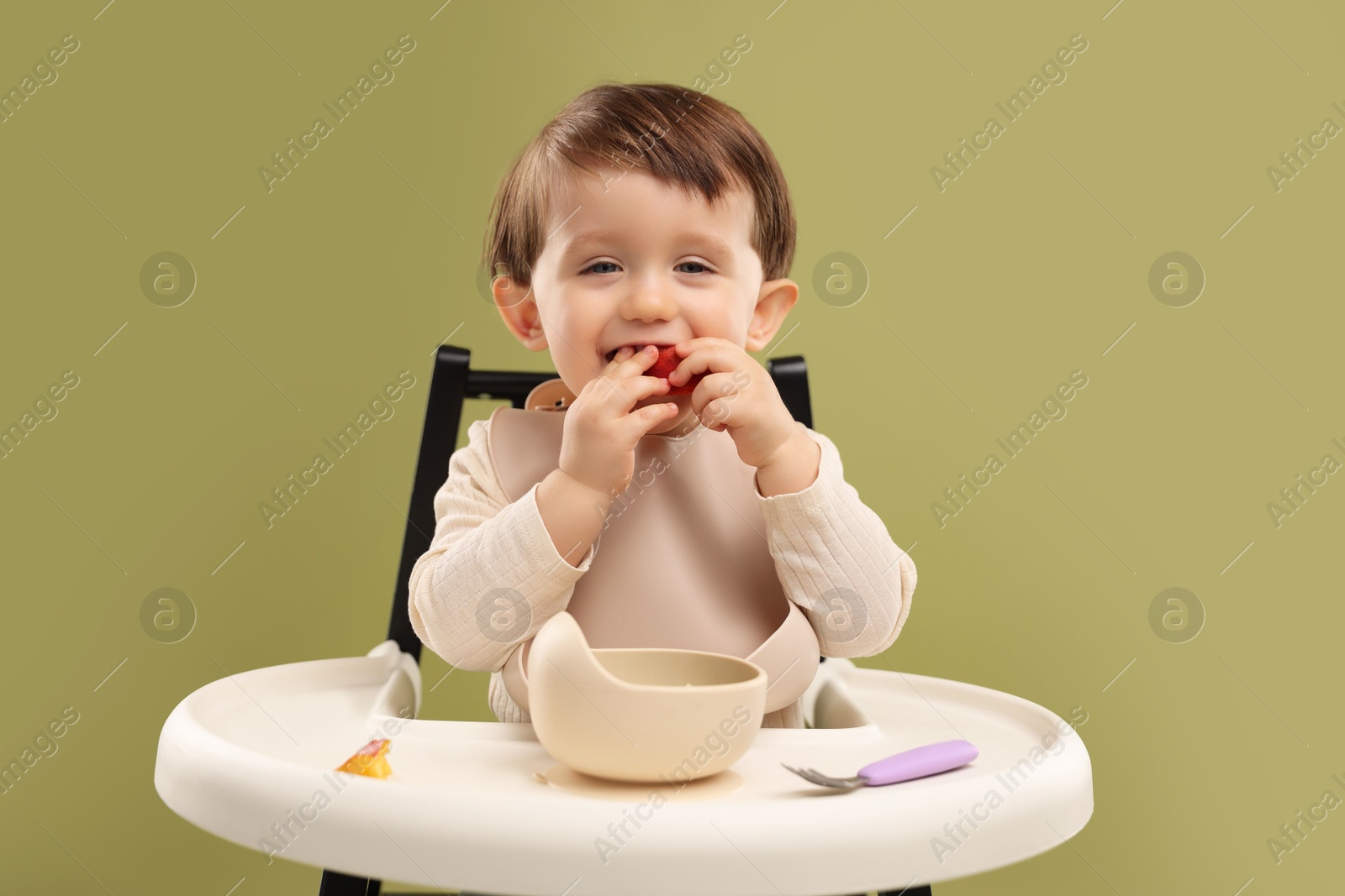 Photo of Cute little baby eating healthy food from bowl in high chair on olive background