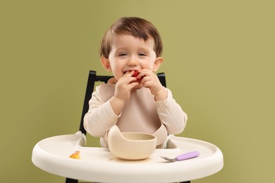 Cute little baby eating healthy food from bowl in high chair on olive background