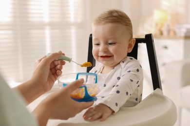 Photo of Mother feeding her cute little baby in high chair at home, closeup