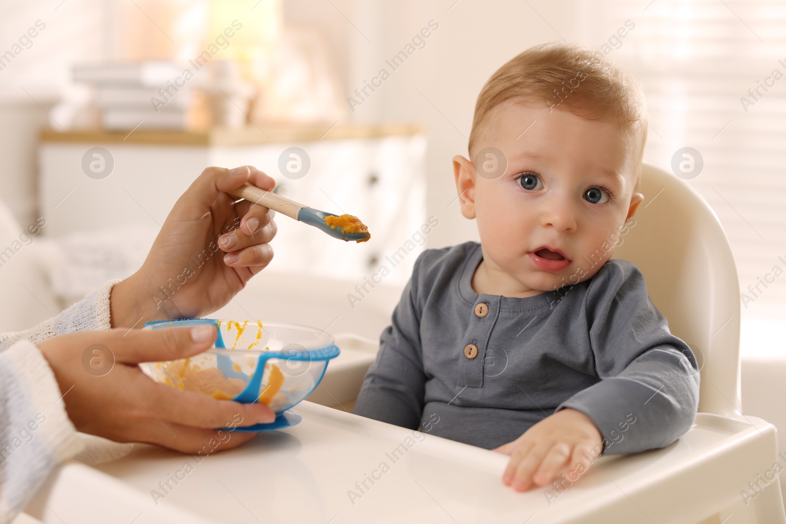 Photo of Mother feeding her cute little baby in high chair at home, closeup