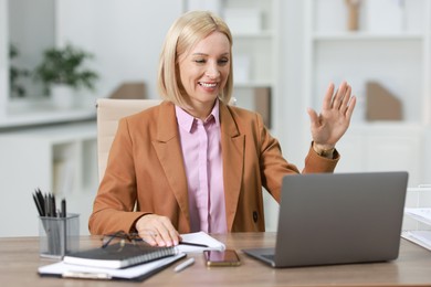 Photo of Smiling middle aged woman having videochat by laptop at table in office