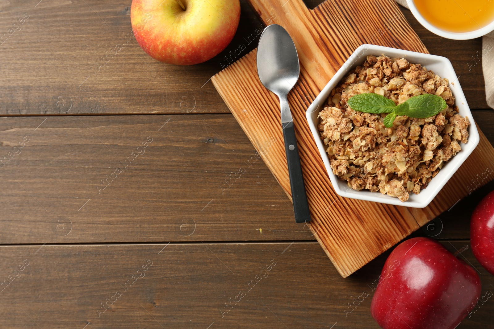 Photo of Tasty apple crisp in bowl, fresh fruits, honey and spoon on wooden table, flat lay. Space for text