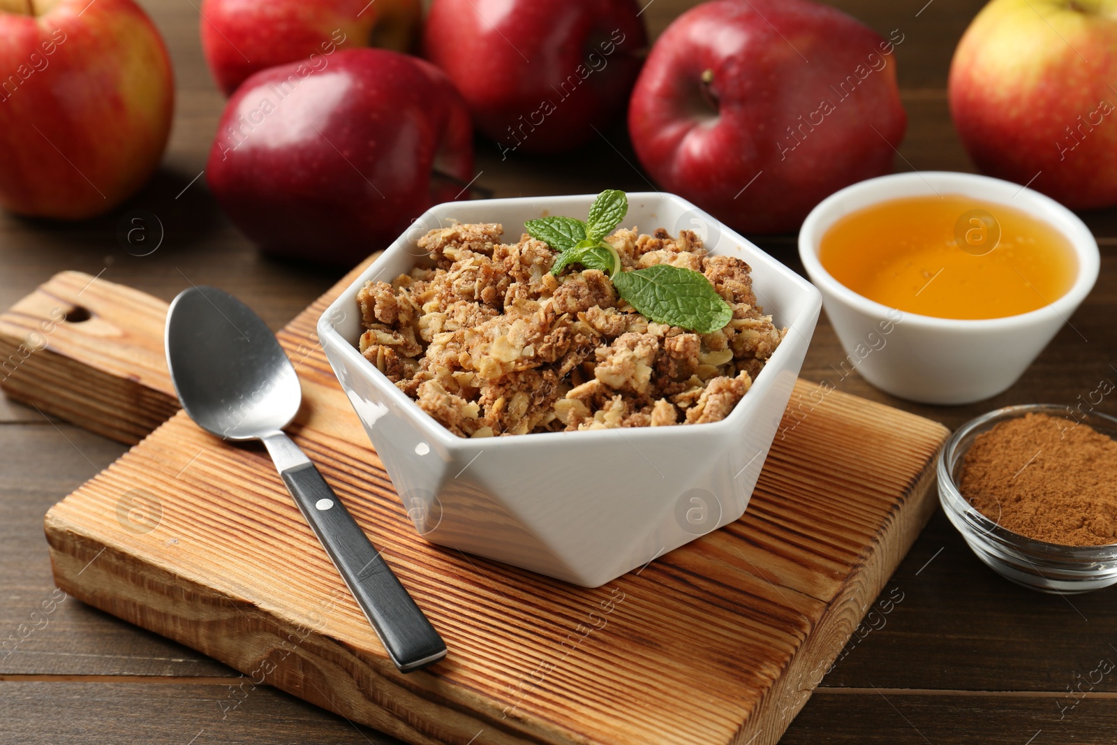 Photo of Tasty apple crisp in bowl, ingredients and spoon on wooden table, closeup