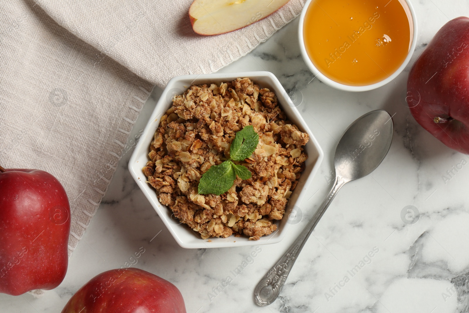 Photo of Tasty apple crisp in bowl, fresh fruits, honey and spoon on white marble table, flat lay