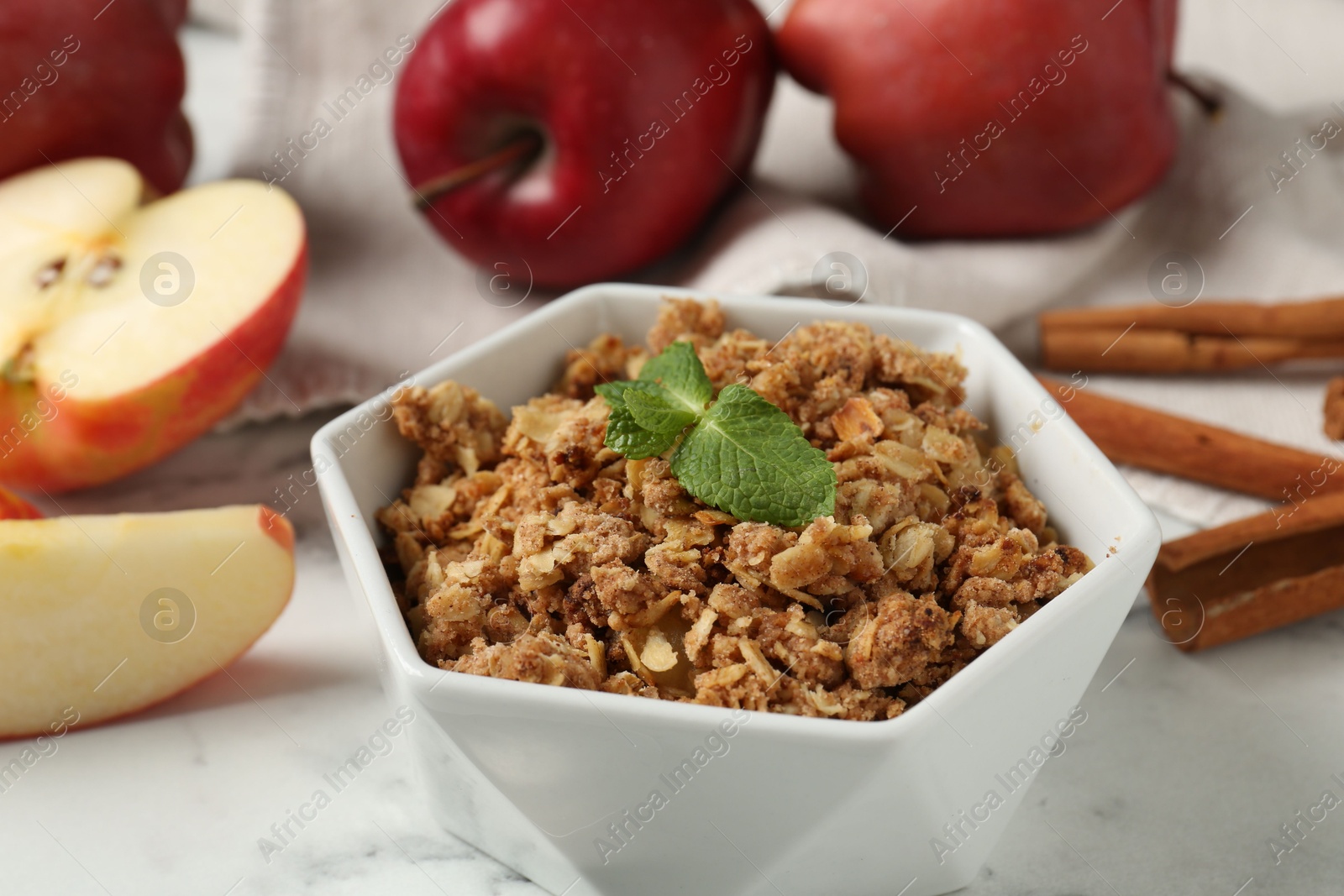 Photo of Tasty apple crisp in bowl, fresh fruits and cinnamon on white marble table, closeup