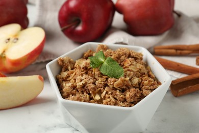 Photo of Tasty apple crisp in bowl, fresh fruits and cinnamon on white marble table, closeup