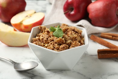 Tasty apple crisp in bowl, ingredients and spoon on white marble table, closeup