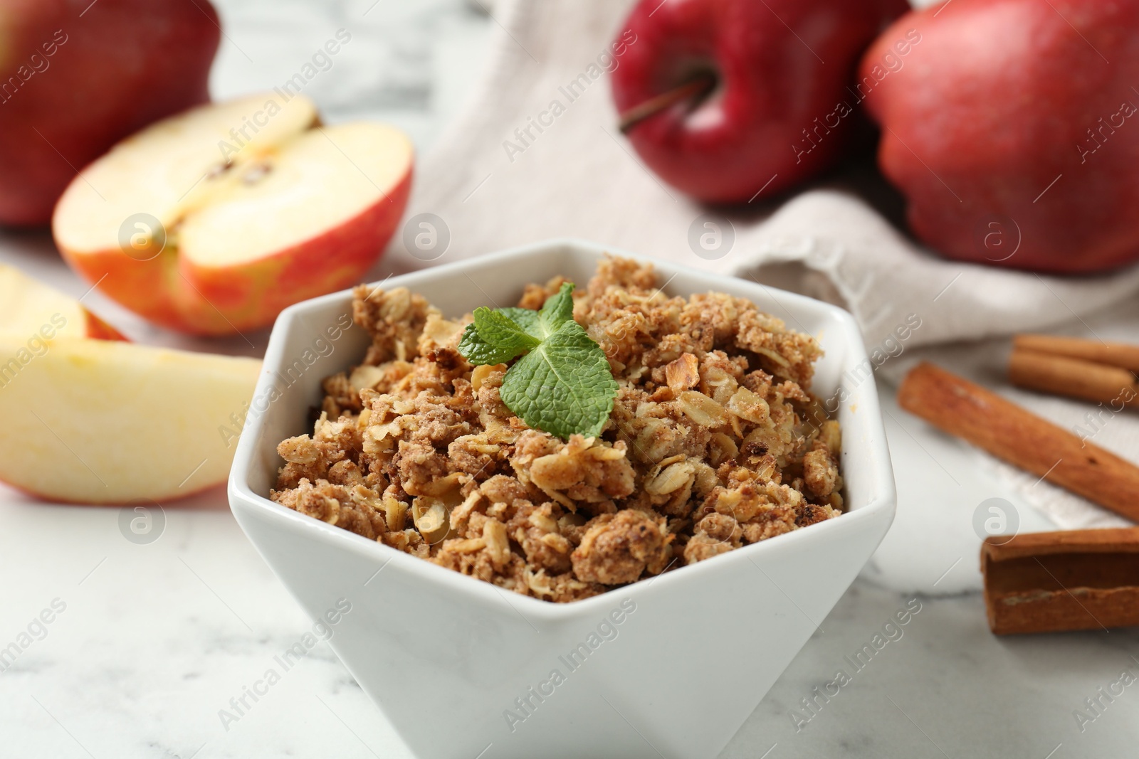 Photo of Tasty apple crisp in bowl, fresh fruits and cinnamon on white marble table, closeup