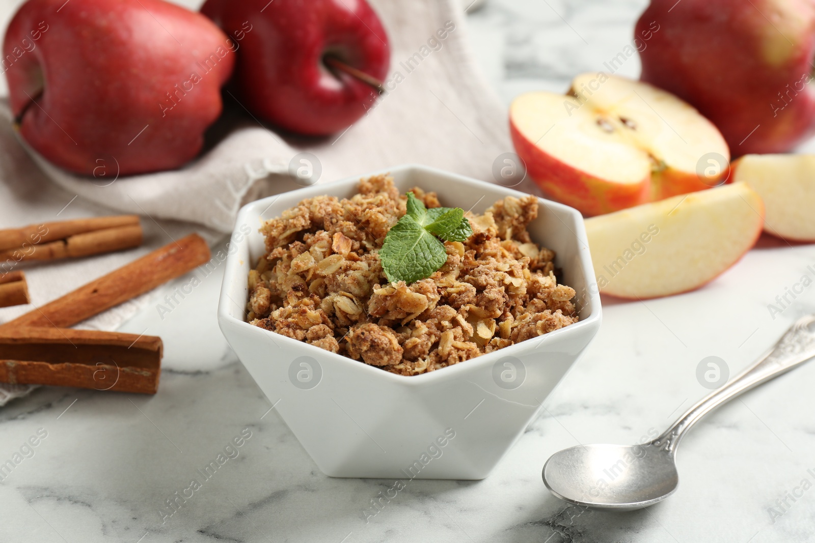 Photo of Tasty apple crisp in bowl, ingredients and spoon on white marble table, closeup