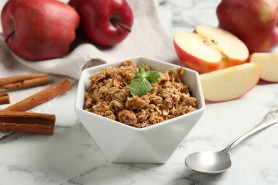 Tasty apple crisp in bowl, ingredients and spoon on white marble table, closeup