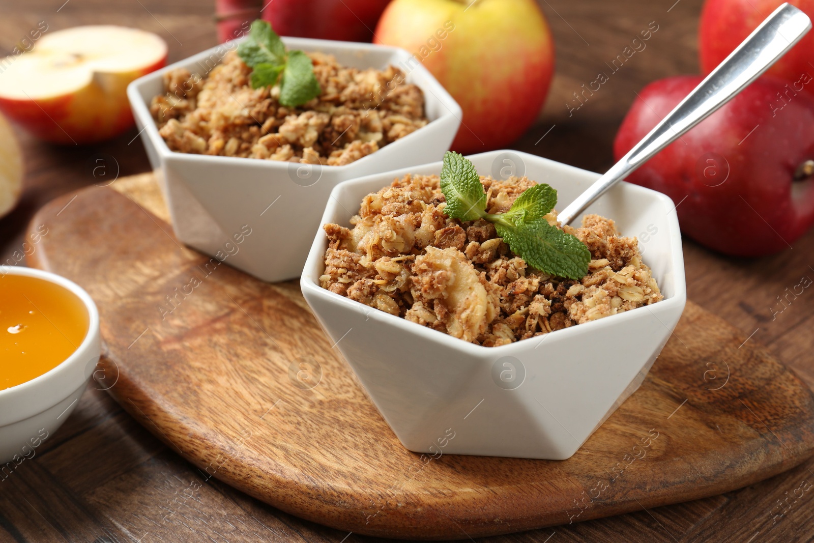 Photo of Tasty apple crisp in bowls, fresh fruits, honey and spoon on wooden table, closeup