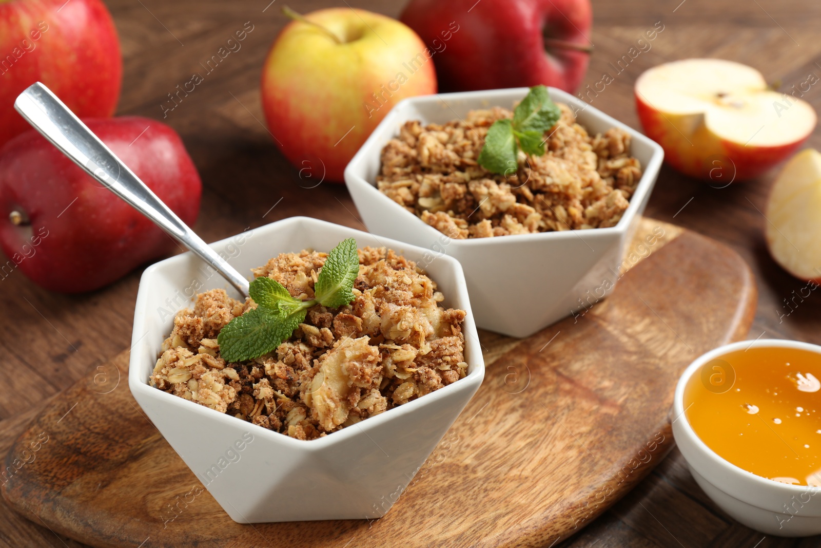 Photo of Tasty apple crisp in bowls, fresh fruits, honey and spoon on wooden table, closeup