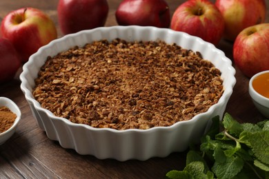 Tasty apple crisp in baking dish and ingredients on wooden table, closeup