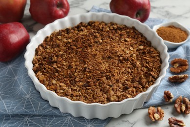 Photo of Tasty apple crisp in baking dish, fresh fruits, cinnamon and walnuts on white marble table, closeup