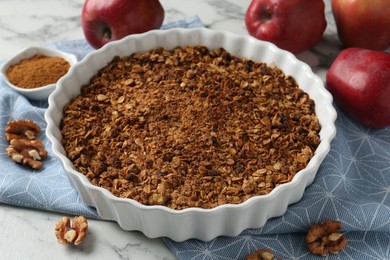 Photo of Tasty apple crisp in baking dish, fresh fruits, cinnamon and walnuts on white marble table, closeup