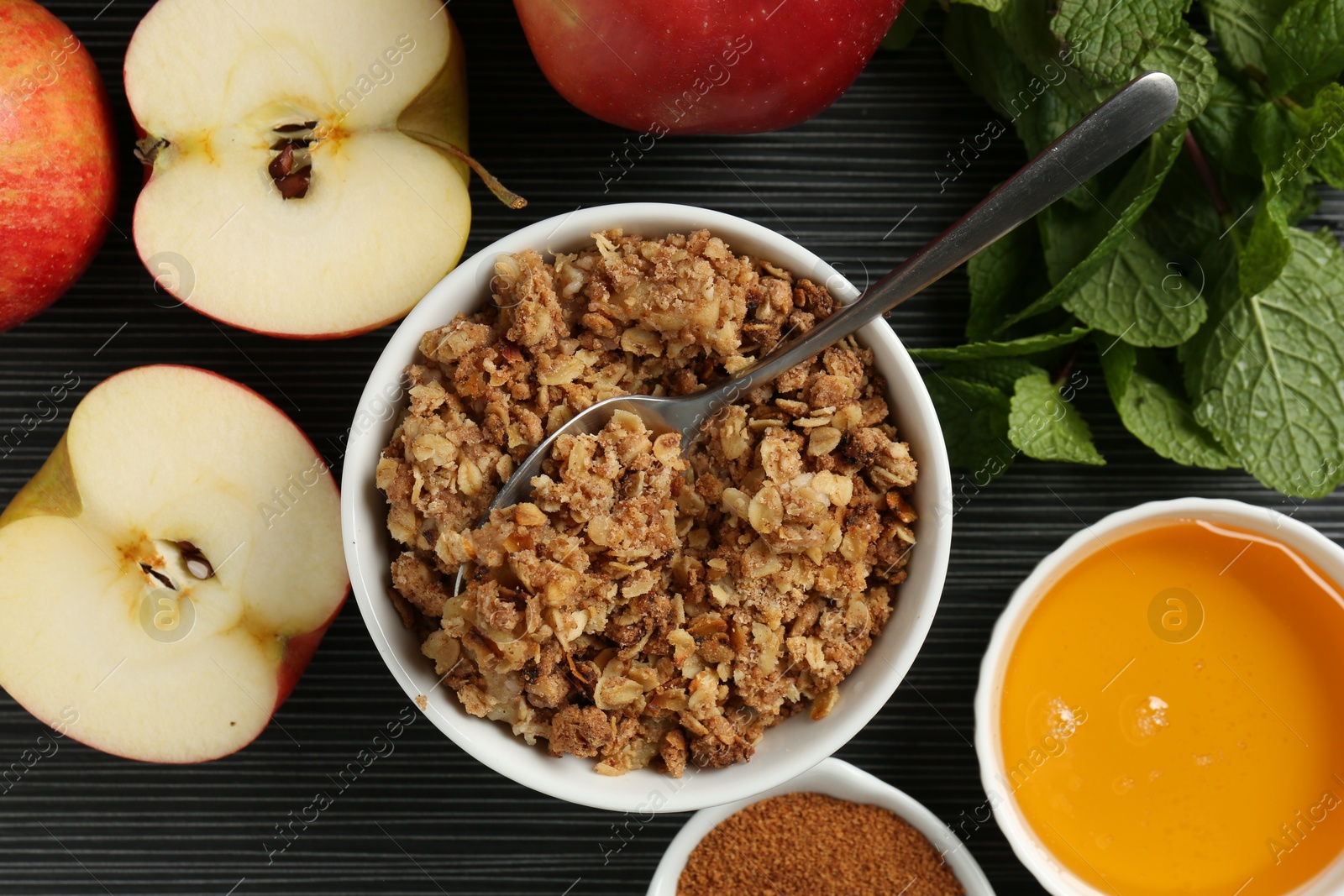 Photo of Delicious apple crisp in bowl, fresh fruits, cinnamon and mint on black textured table, flat lay