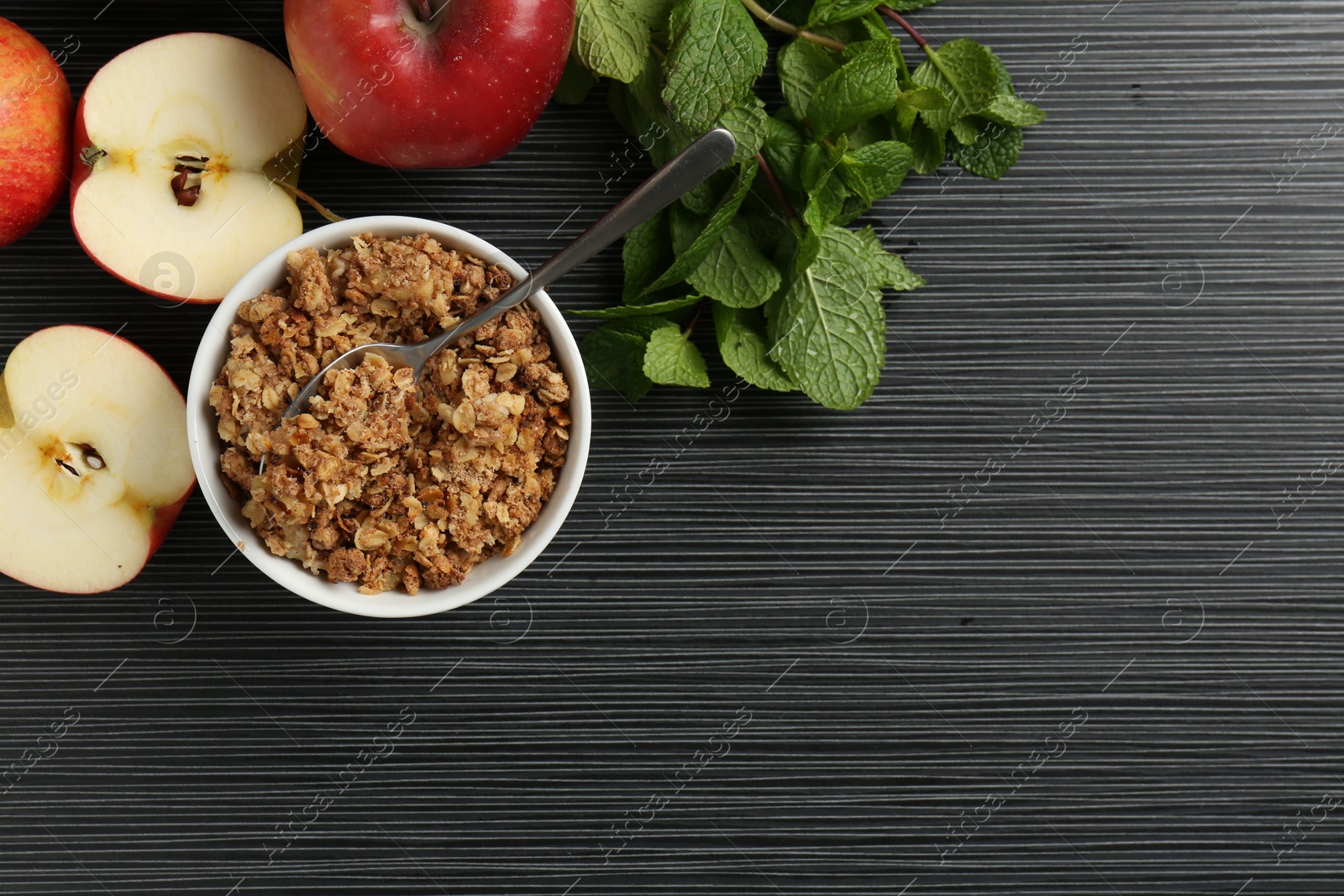 Photo of Delicious apple crisp in bowl, fresh fruits and mint on black textured table, flat lay. Space for text