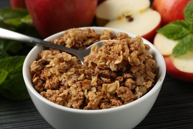 Photo of Delicious apple crisp in bowl, fresh fruits and mint on black textured table, closeup