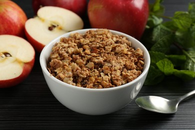 Photo of Delicious apple crisp in bowl, fresh fruits and mint on black textured table, closeup