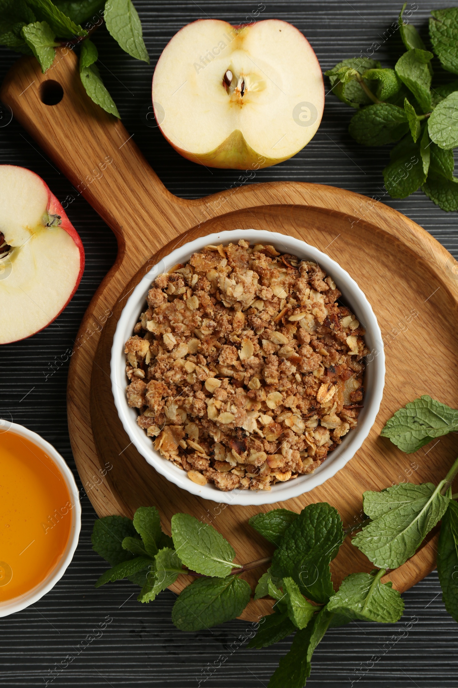 Photo of Delicious apple crisp in bowl, fresh fruits, honey and mint on black textured table, flat lay