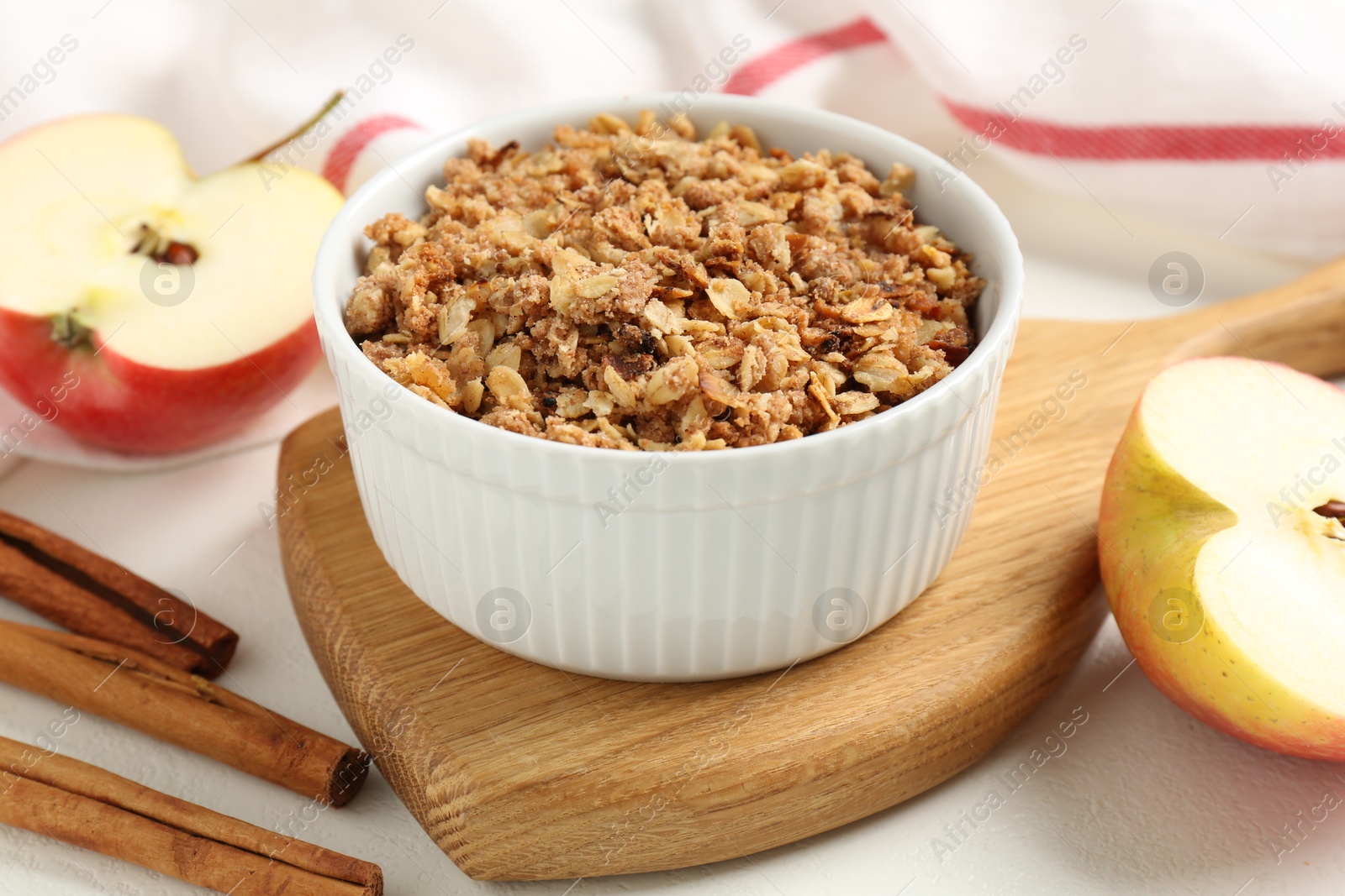 Photo of Delicious apple crisp in bowl, fresh fruits and cinnamon sticks on white table, closeup