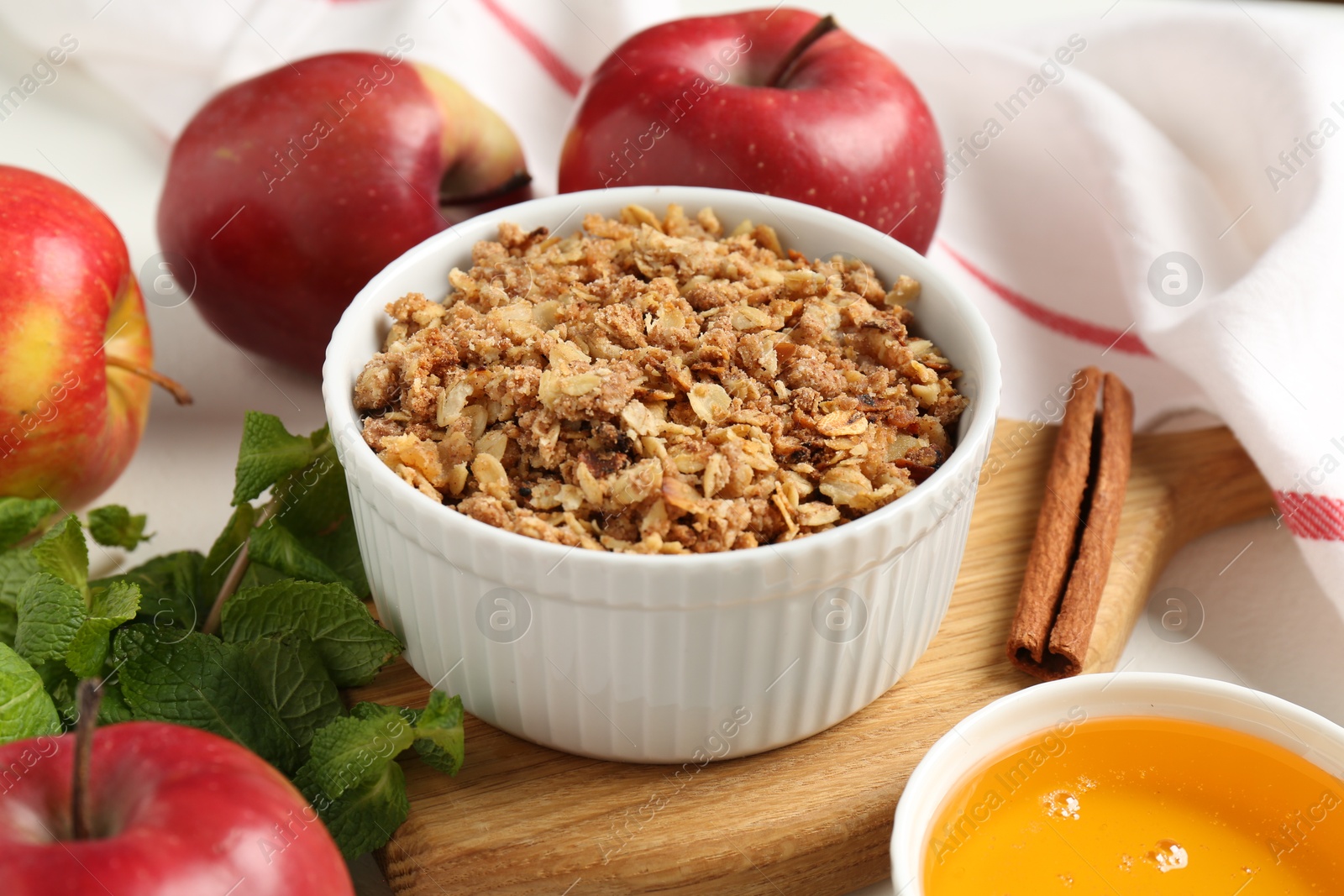 Photo of Delicious apple crisp in bowl, fresh fruits, mint, honey and cinnamon stick on table, closeup