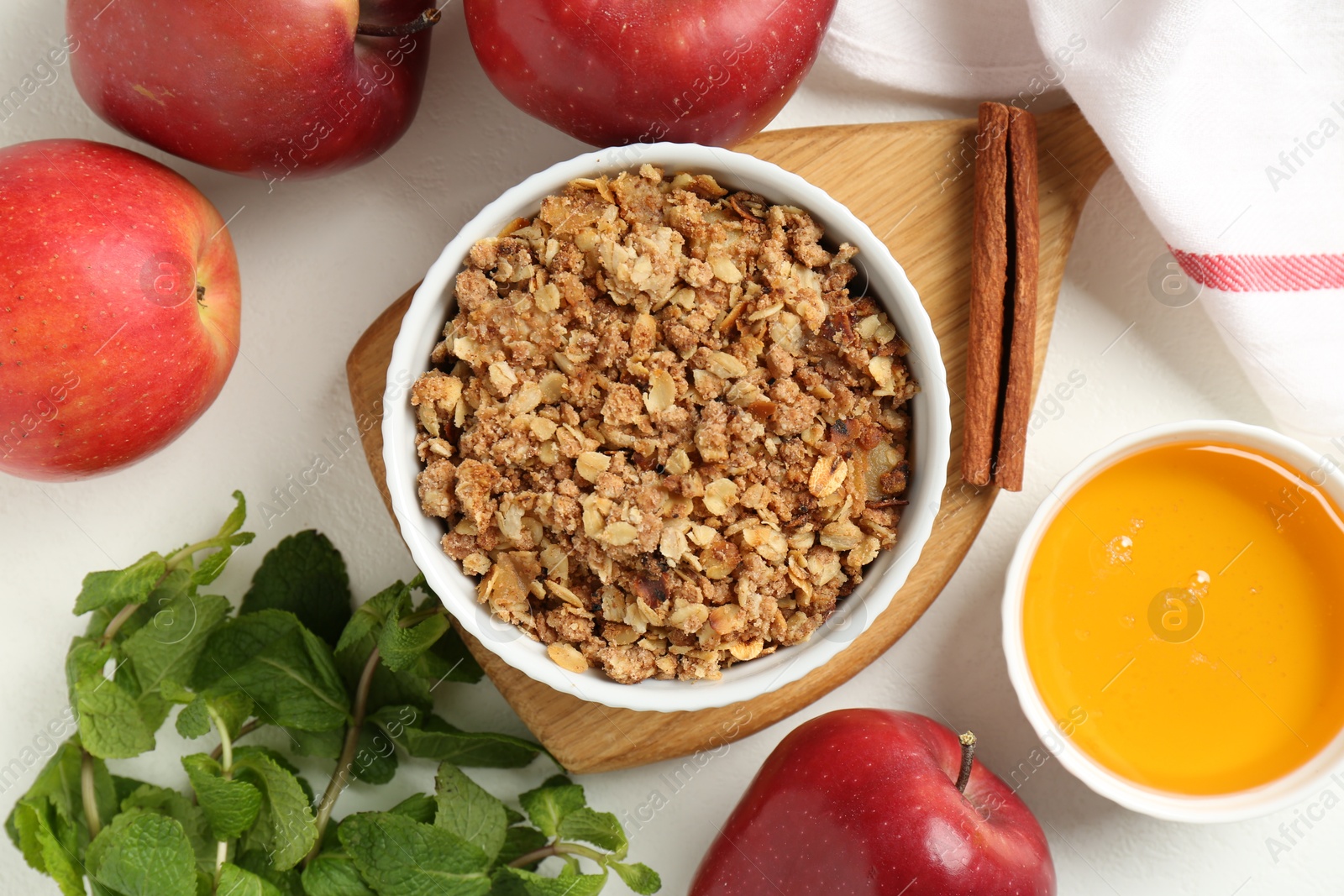 Photo of Delicious apple crisp in bowl, fresh fruits, mint, honey and cinnamon stick on white table, flat lay