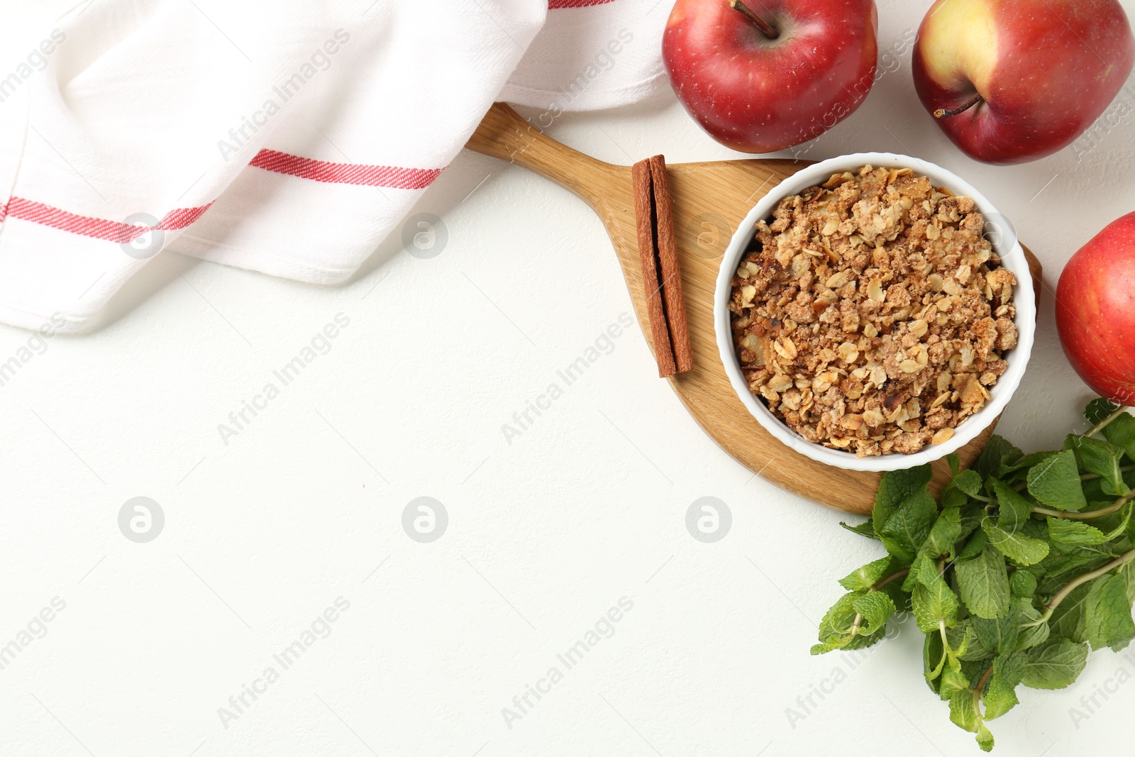 Photo of Delicious apple crisp in bowl, fresh fruits, mint and cinnamon stick on white table, flat lay. Space for text