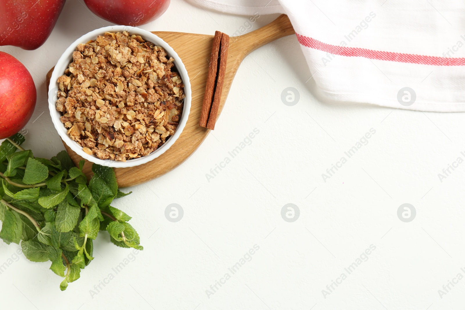 Photo of Delicious apple crisp in bowl, fresh fruits, mint and cinnamon stick on white table, flat lay. Space for text