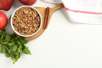 Photo of Delicious apple crisp in bowl, fresh fruits, mint and cinnamon stick on white table, flat lay. Space for text