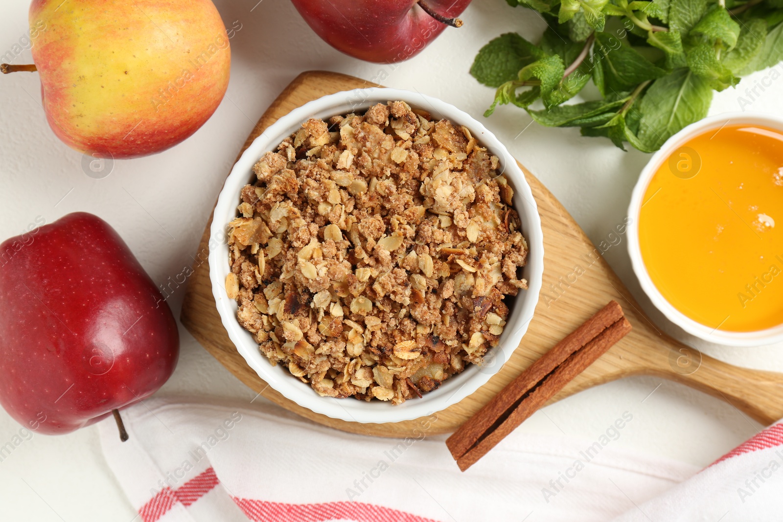 Photo of Delicious apple crisp in bowl, fresh fruits, mint, honey and cinnamon stick on white table, flat lay