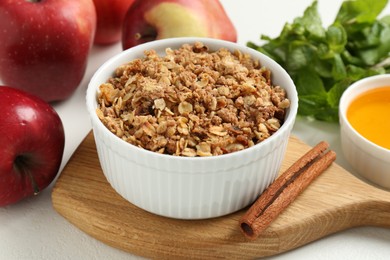 Photo of Delicious apple crisp in bowl, fresh fruits, cinnamon stick, honey and mint on white table, closeup