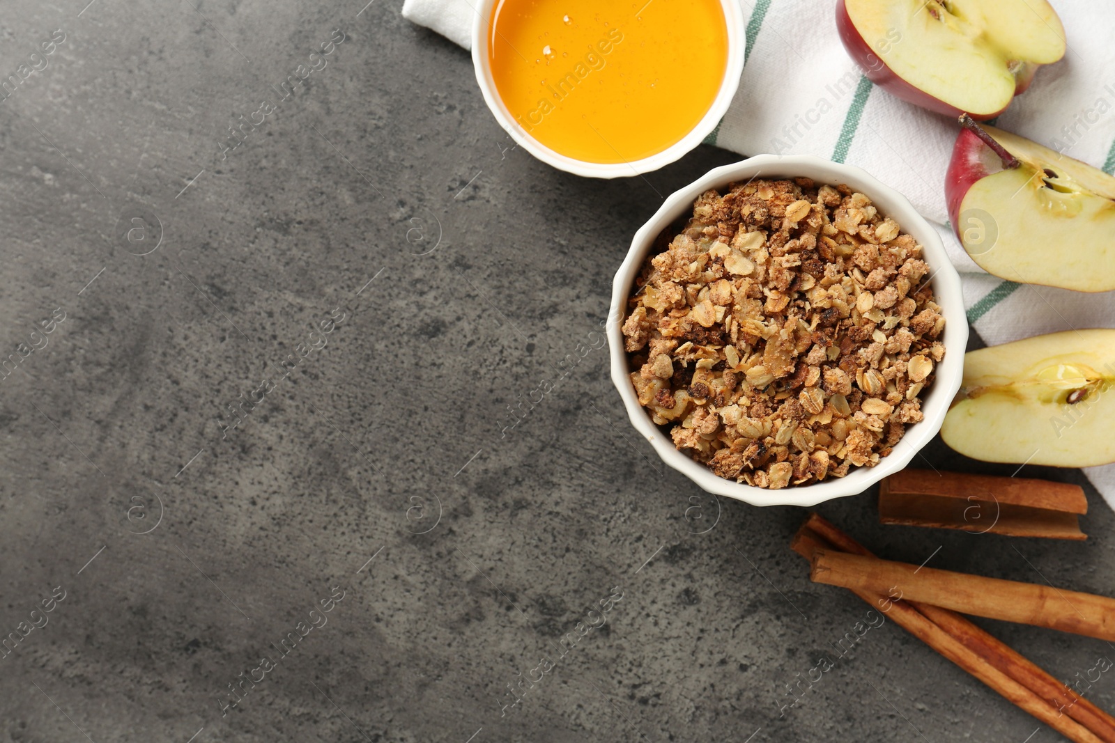 Photo of Delicious apple crisp in bowl, fresh fruits, cinnamon sticks and honey on grey table, flat lay. Space for text