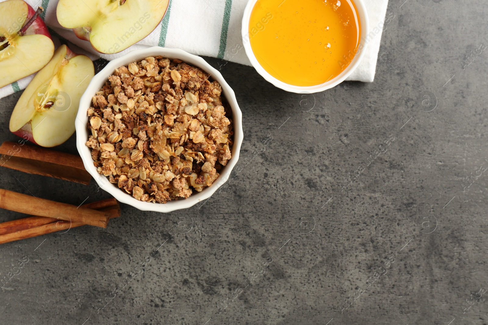 Photo of Delicious apple crisp in bowl, fresh fruits, cinnamon sticks and honey on grey table, flat lay. Space for text