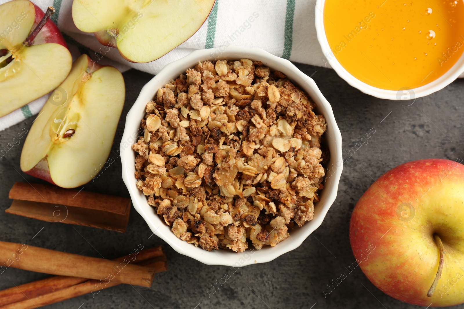 Photo of Delicious apple crisp in bowl, fresh fruits, cinnamon sticks and honey on grey table, flat lay