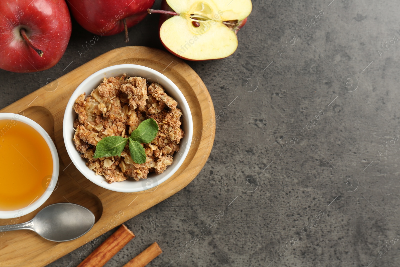 Photo of Delicious apple crisp in bowl, fresh fruits, cinnamon, honey and mint on grey table, flat lay. Space for text