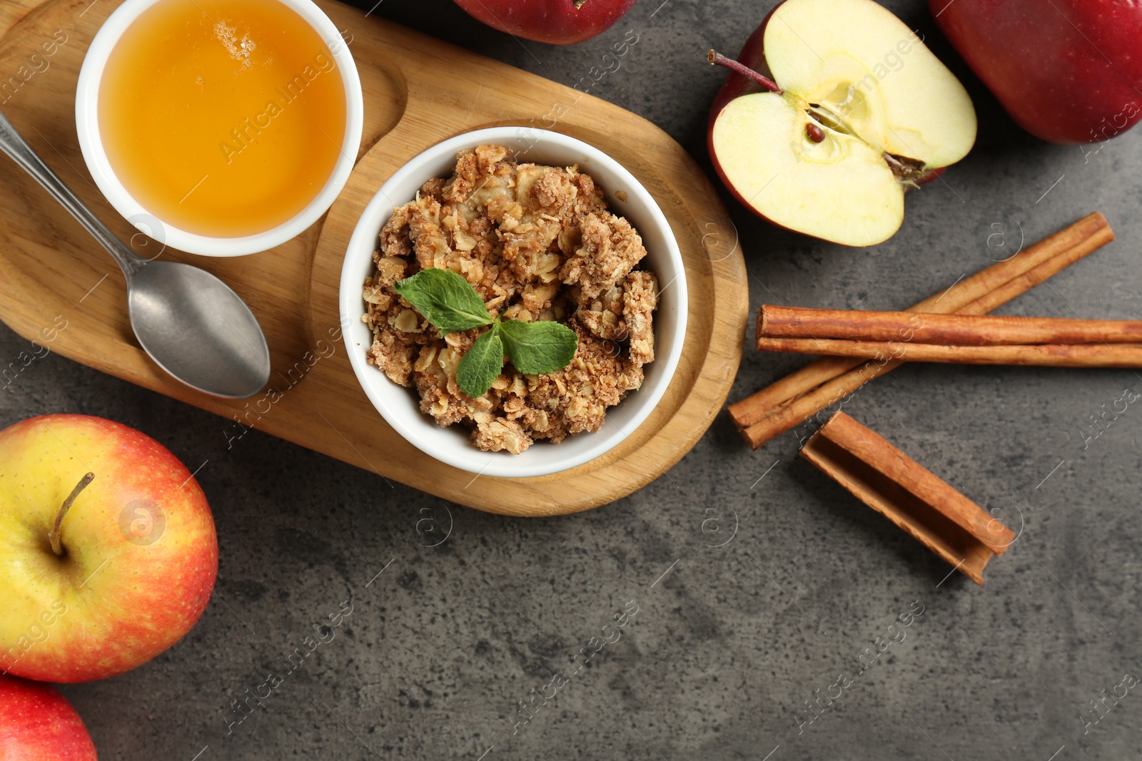 Photo of Delicious apple crisp in bowl, fresh fruits, cinnamon sticks, honey and mint on grey table, flat lay