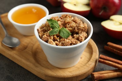 Photo of Delicious apple crisp in bowl, fresh fruits, cinnamon sticks, honey and mint on grey table, closeup