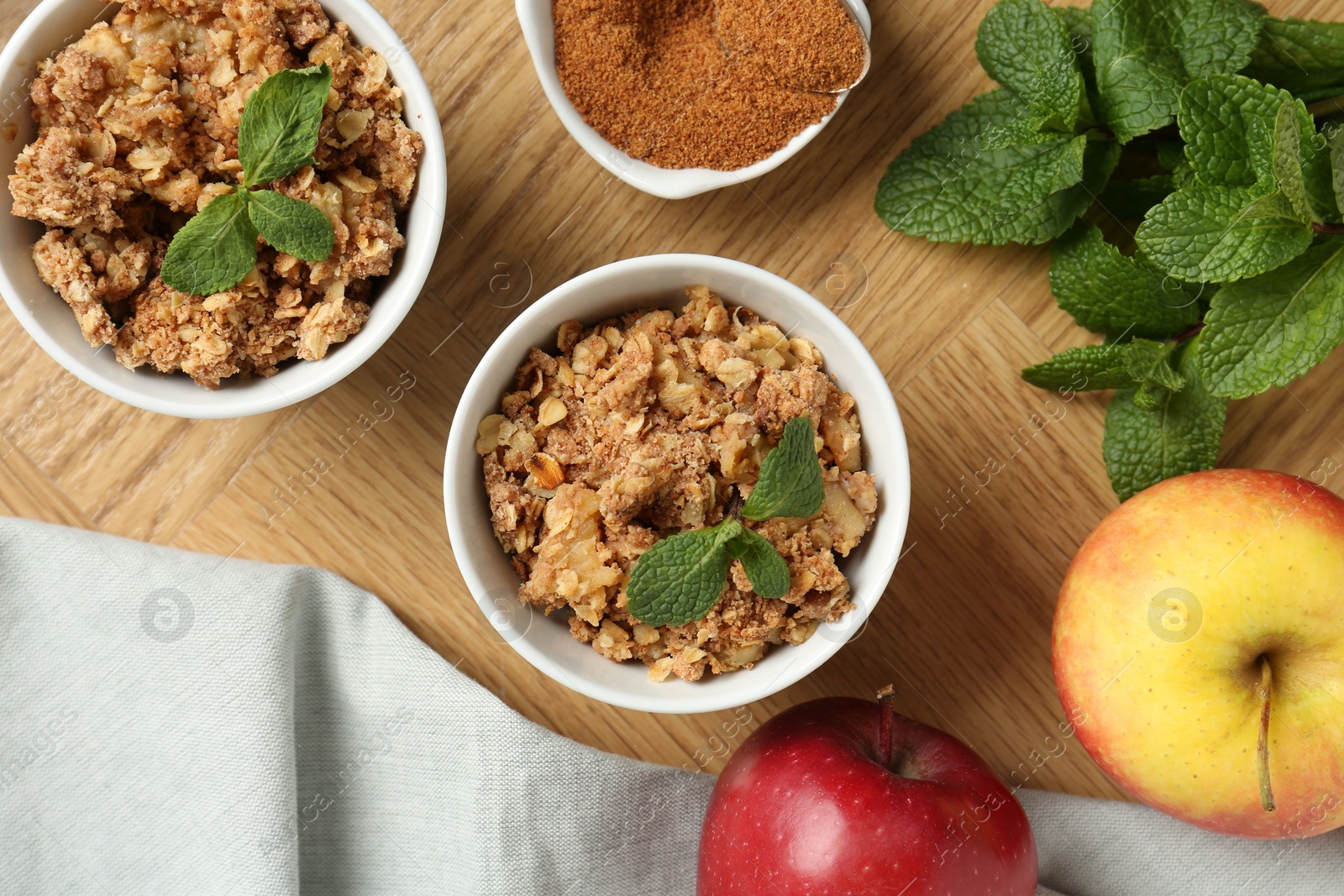 Photo of Delicious apple crisp in bowls, fresh fruits, mint and cinnamon on wooden table, flat lay